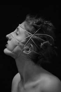 A black and white photograph of a young woman with rubber bands around her face distorting her features