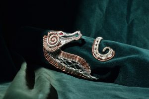 A cooper and silver seahorse shaped bracelet with a red bead eye. Presented on a green velvet background. 