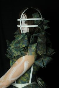 A photograph of a woman with a painted white face wearing a cage like head piece. The garment she wears is made of a series of folded paper shapes each with green botanical prints on them. 