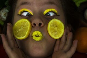 A landscape photograph of a girl with bright yellow eyeshadow, lemon slices on her cheeks and yellow lipstick. She is cross eyed and pursing her lips. 