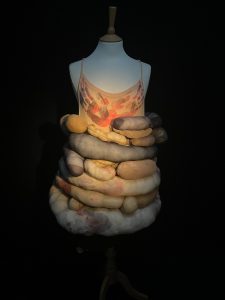 A dress on a mannequin. The top half of the dress is pink with thin straps over the chest area there are red and black hand prints.  