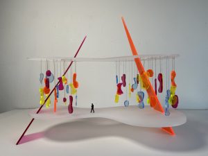 A plastic diorama of a curved elevated space with smaller orange, yellow, blue and purple curved shapes hanging overhead a tiny model of a person. 