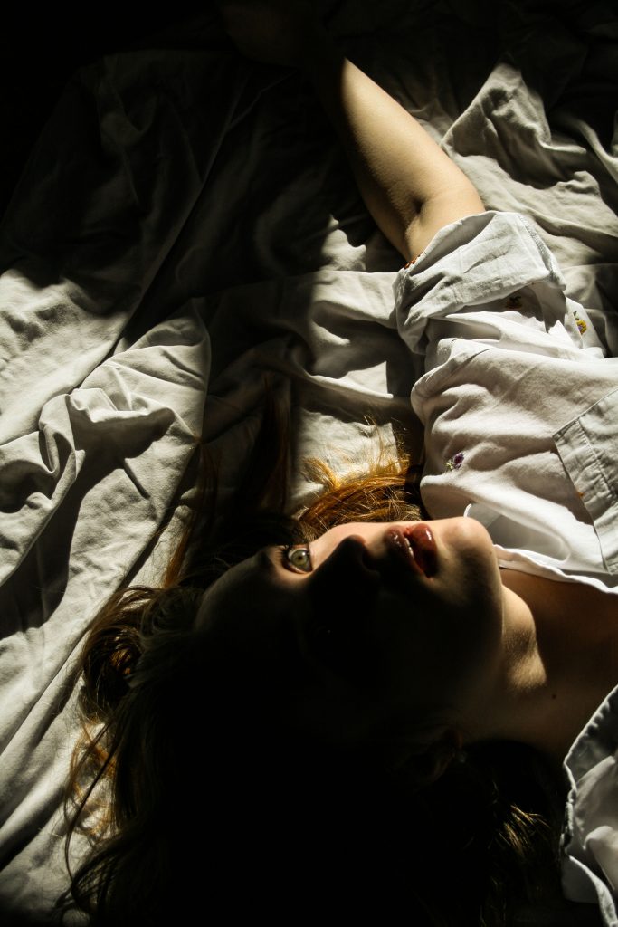 A photograph of a young woman laying down on a sheet. Only her head and arm are in view but half her face is obscured by a dark shadow.  