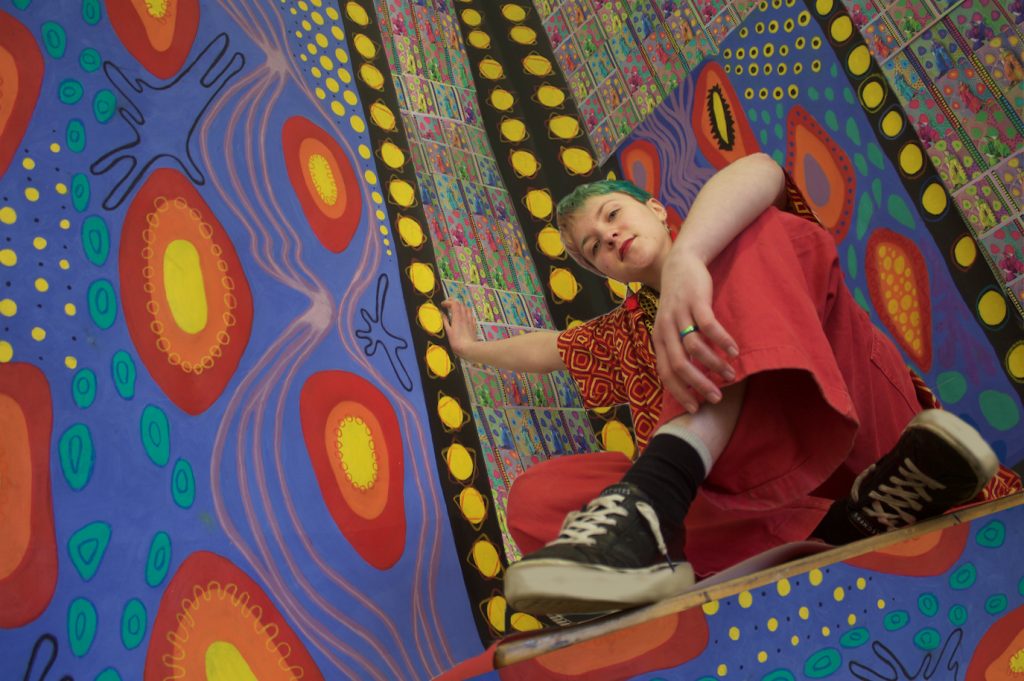 A photograph of a young person with short blue and blond hair they are wearing bright red and yellow clothing. They are sitting in a brightly patterned space. 