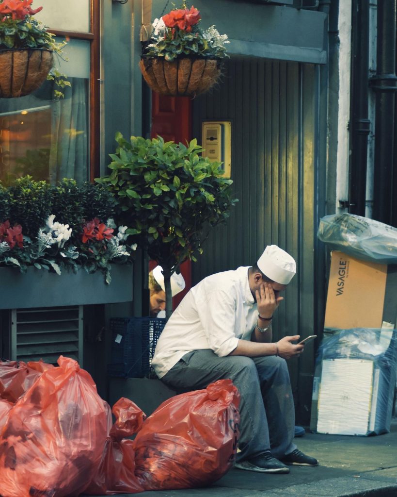 A photograph of a couple chefs on their break outside an establishment both are looking at their phones. They are surrounded by hanging flowers on side of the image and the other rubbish bags and carboard. 