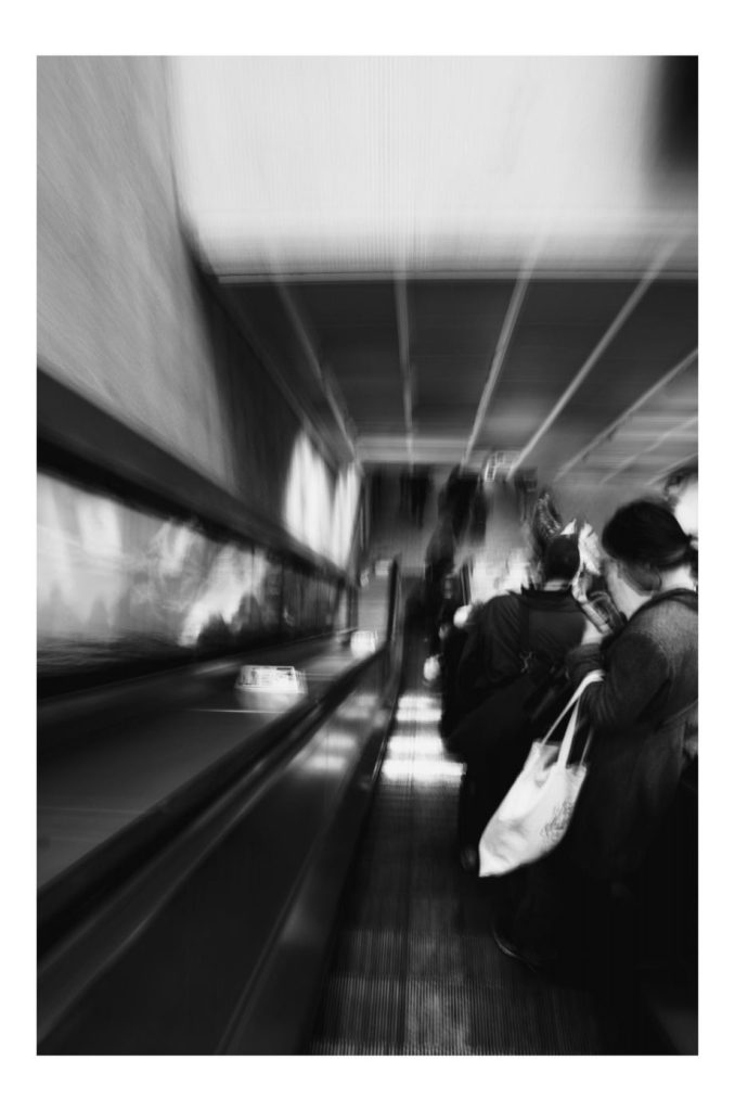 A black and white photograph of an London tube escalator. There are people standing to one side. The exposure is blurry.  