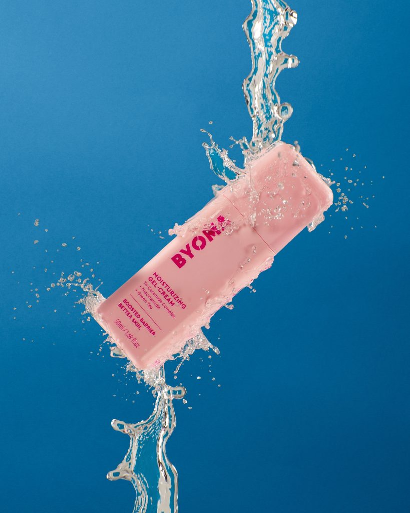 A photograph of a pink moisturiser bottle. It is suspended by water jets from the bottom and the top. 