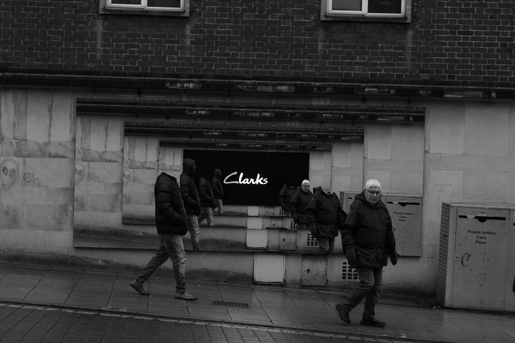 A digitally altered photograph of two men walking past a Clarks shop sign. The photograph has been repeated where the sign was and getting small each time, the men's heads have been removed. 