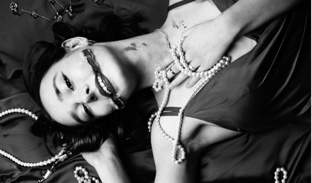 A photograph of a young woman laying down, she is looking at the viewer and clutching a pearl necklace. Her mouth has been cut across, on neck and chest is flecks of blood. 